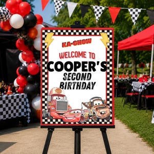 Puede incluir: Un letrero de cumplea&ntilde;os rectangular sobre un caballete negro con un borde a cuadros, rayos amarillos y el texto KA-CHOW WELCOME TO COOPER'S SECOND BIRTHDAY. El letrero muestra ilustraciones de tres coches de carreras de dibujos en rojo y marr&oacute;n.