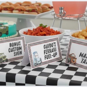 May include: Three white rectangular food tent cards with black and white checkered borders. The cards feature cartoon car characters and text reading Bonus Custom Sign, Guido's Ferrari Fuel-Up, and Tow Mater's Taters in a bold black font.