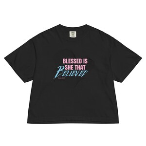 Op de afbeelding: Zwart T-shirt met de tekst "BLESSED IS SHE THAT BELIEVED" in roze en blauw. Het shirt heeft een relaxte pasvorm en korte mouwen. De tekst is in een gestileerd lettertype.