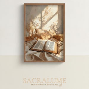 May include: A framed wall art print featuring an illustration of an open book resting on rumpled white bed sheets. Sunlight streams through a window, casting shadows across pillows and a small vase with dried grass. The text SACRALUME Downloadable Christian Art is printed below the frame.