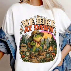 Può includere: Una t-shirt bianca con la grafica di una rana verde che indossa un cappello da escursionismo, uno zaino e stivali. La rana tiene un bastone da passeggio vicino a un falò e una tazza. Il testo recita WE HIKE AT DAWN, WILD PLACES, GOOD PACE, CLEAR MIND e TAKE THE SCENIC ROUTE.