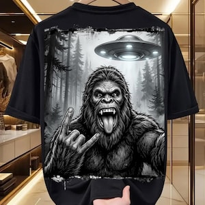 May include: Black t-shirt featuring a graphic of a gorilla making a rock and roll hand gesture, with a forest backdrop and a UFO. The gorilla is depicted with its tongue out. The design is in black and white.