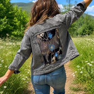 May include: A gray acid-wash denim jacket featuring a large rectangular back panel made of sheer black mesh fabric. The mesh panel is decorated with embroidered floral patterns in shades of blue, white, and tan, with long sleeves and buttons.