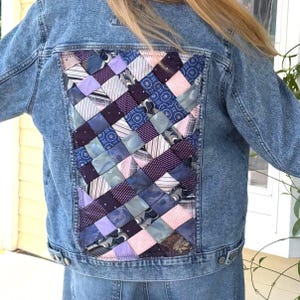 May include: A blue denim jacket featuring a large rectangular back panel composed of woven fabric strips in various shades of purple, blue, pink, and white. The woven section displays a mix of solid colors, stripes, and floral patterns.