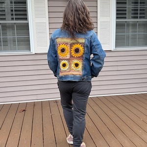 May include: A blue denim jacket featuring a large rectangular crochet panel on the back. The panel consists of four square motifs, each displaying a sunflower design with yellow petals and a dark brown center, set against a light tan background.