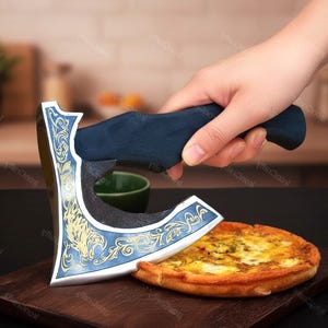 May include: A pizza cutter shaped like a battle axe with a dark blue handle and a silver blade featuring gold-coloured filigree engravings and a wolf head design. The blade is positioned to slice through a round, golden-brown baked pizza.