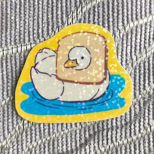 以下が含まれることがあります： 青い水に浮かぶ、トーストを頭にした漫画のアヒルの風変わりなステッカー。ステッカーは黄色の縁取りとキラキラとしたアクセントがあり、遊び心のある目を引くデザインになっています。
