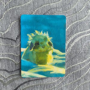 May include: A rectangular card featuring a painted illustration of a small, green, fluffy creature with pointed ears and a spiky crown, set against a vibrant blue background with light yellow highlights on the ground beneath the figure.