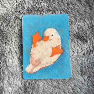 May include: A rectangular card featuring a whimsical painting of a white duck with orange feet and beak against a blue background. The duck is depicted in a playful pose, with its head tilted and wings slightly spread. The artwork has a textured, painted appearance.