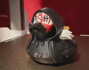 May include: A black rubber duck figurine dressed in a hooded cloak with a white face mask featuring red markings. The duck has a black beak, a necklace with small skull charms, a belt around its waist, and a white ruffled collar with a small orange flower.
