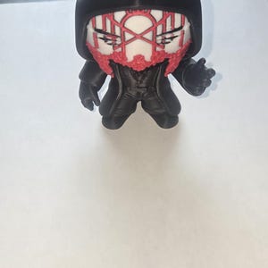 May include: A black figurine with a hooded cloak and a stylized face featuring red accents. The figure has a detailed design, with a textured hood and clothing. The figurine is standing on a white surface.