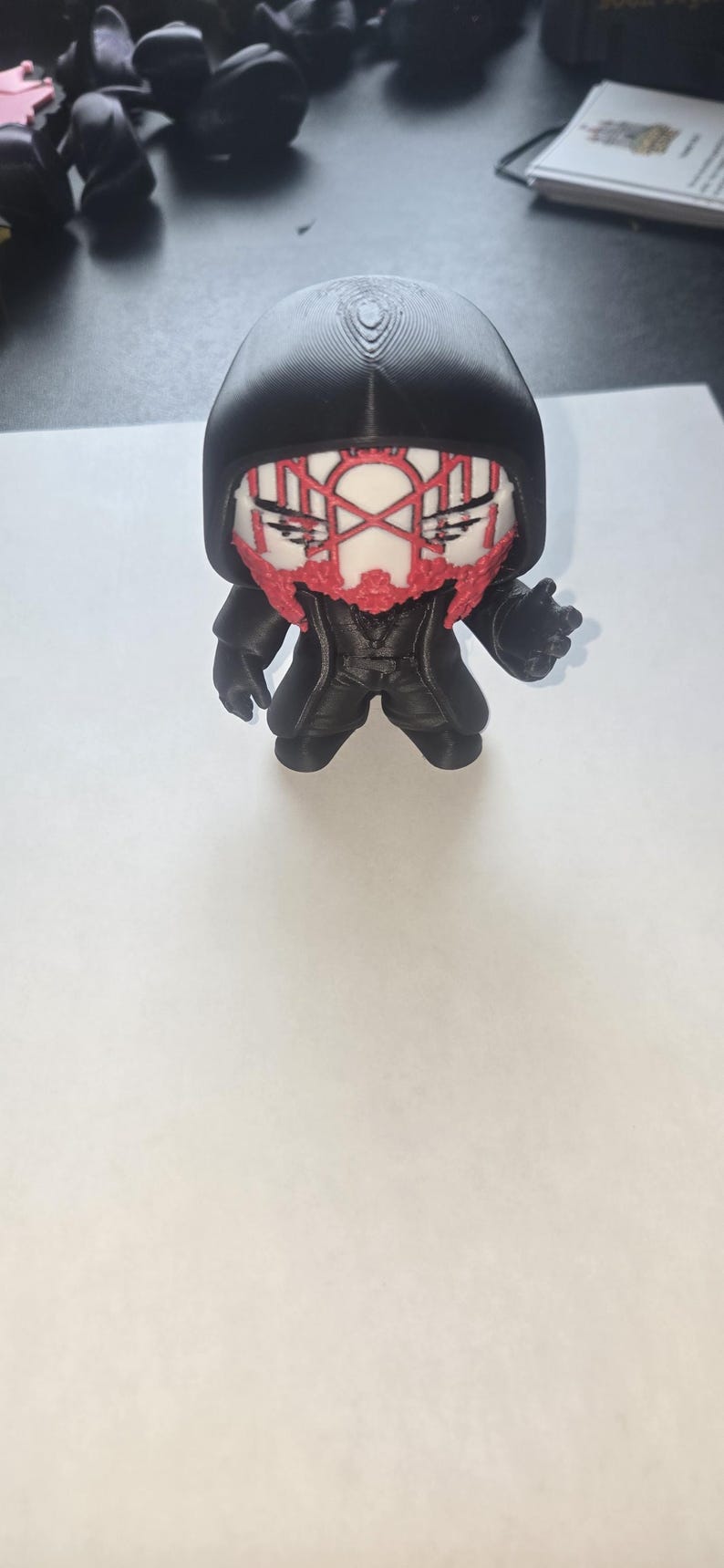 May include: A black figurine with a hooded cloak and a stylized face featuring red accents. The figure has a detailed design, with a textured hood and clothing. The figurine is standing on a white surface.