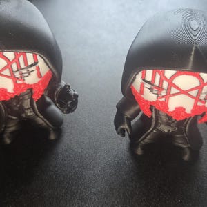 May include: Two black figurines with a stylized design. Each figure has a black hood, a red and white face design, and a black coat. The figures are small and appear to be 3D printed.