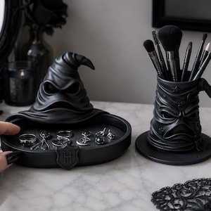 May include: A matte black resin jewellery tray shaped like a wizard hat with a shield featuring the letter H, containing several silver rings. Beside it sits a matching black wizard hat cup filled with various makeup brushes on a white surface.