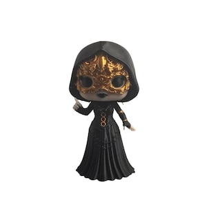 May include: A stylized collectible figurine featuring a character wearing a black hooded cloak and a detailed gold mask. The figure has a long black dress with vertical pleats and a gold circular emblem on the chest. The character has dark lips.