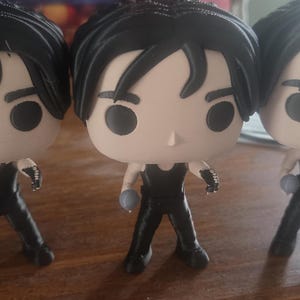 May include: Three Funko Pop vinyl figures of a character with black hair and a black outfit. Each figure has a light skin tone and is holding a small, gray object. The figures are standing on a wooden surface.