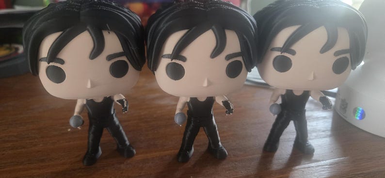 May include: Three Funko Pop vinyl figures of a character with black hair and a black outfit. Each figure has a light skin tone and is holding a small, gray object. The figures are standing on a wooden surface.