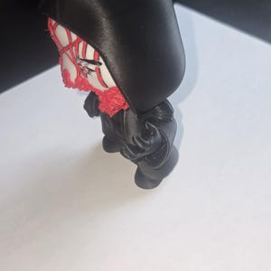 May include: A black figurine with a hood, featuring a white face with red accents. The figure has a red beard and is posed in a standing position. The figurine is on a white surface.