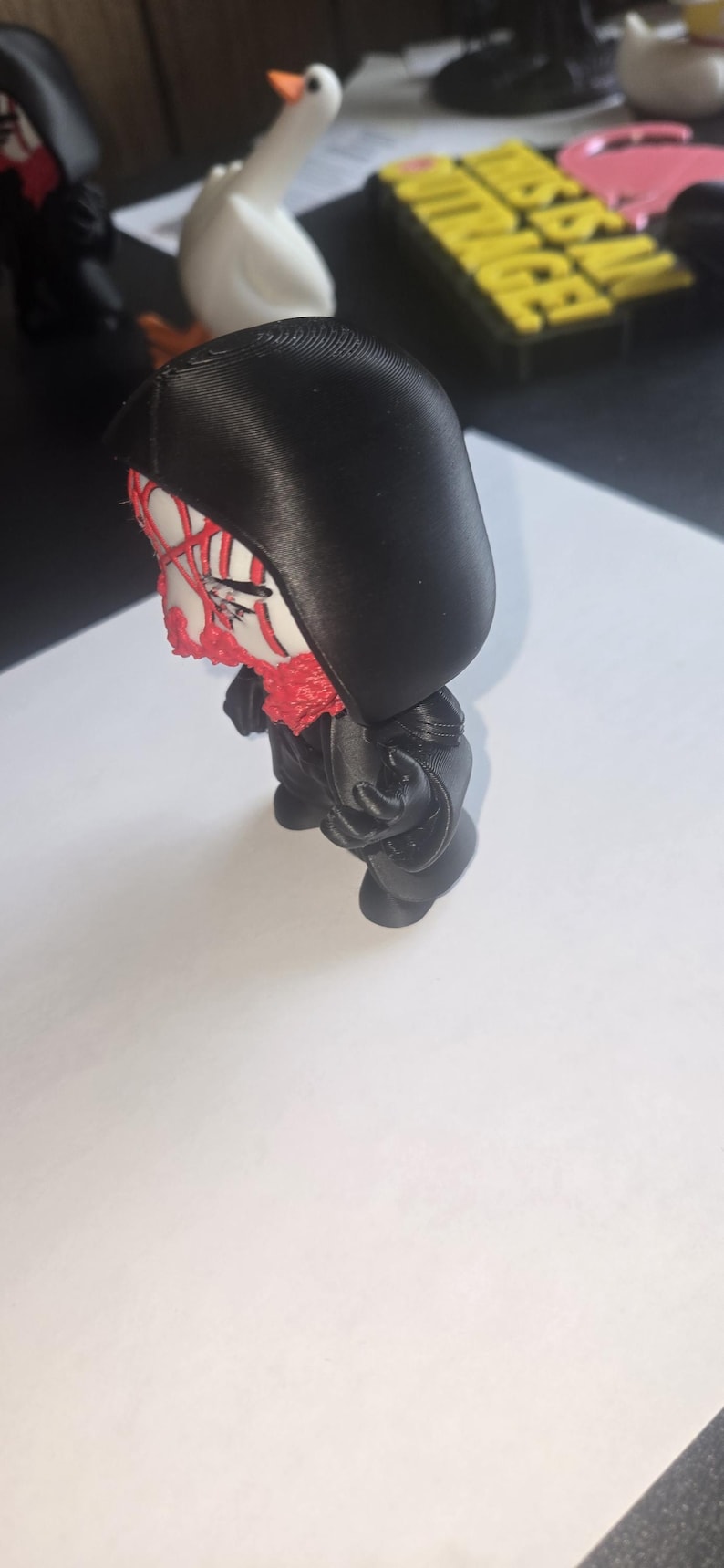 May include: A black figurine with a hood, featuring a white face with red accents. The figure has a red beard and is posed in a standing position. The figurine is on a white surface.