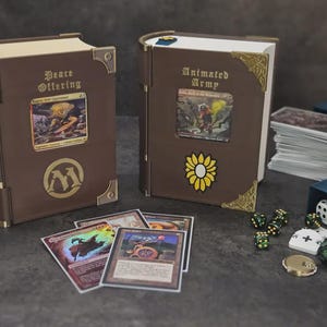 May include: Two brown book-shaped card holders with gold accents and card art. One has a sunflower sticker, the other a gold "M" logo. Several trading cards, dice, and a coin are scattered around the holders.