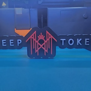 May include: A black sign featuring the text SLEEP TOKEN in white capital letters, separated by a central red geometric logo with a sun-like crest. The sign has a scalloped edge design and is positioned against a dark, industrial-style background.