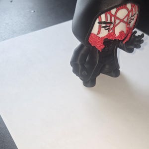 May include: A black figurine of a hooded figure with a red and white face mask. The figure is standing on a white surface. The figurine has a detailed design and is likely 3D printed.