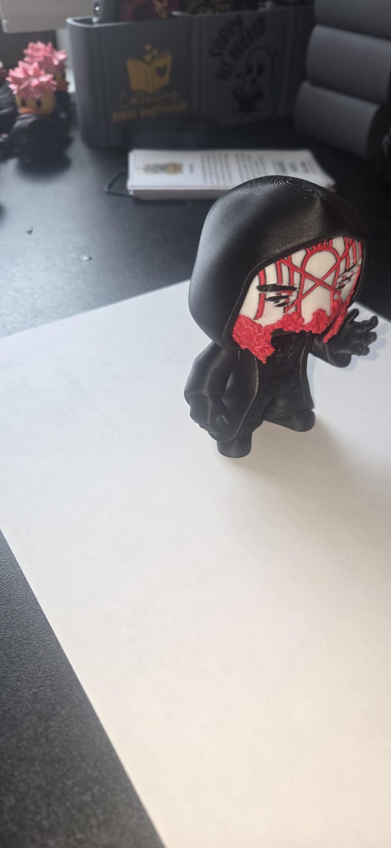 May include: A black figurine of a hooded figure with a red and white face mask. The figure is standing on a white surface. The figurine has a detailed design and is likely 3D printed.