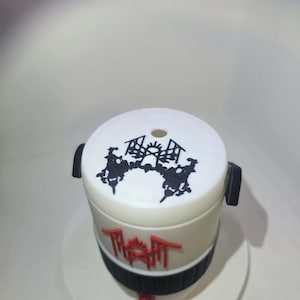 May include: A white cylindrical container featuring a black graphic design on the top lid and a red graphic logo on the side. The base of the container is wrapped in a black ribbed band, and a small red component is attached to the bottom center.