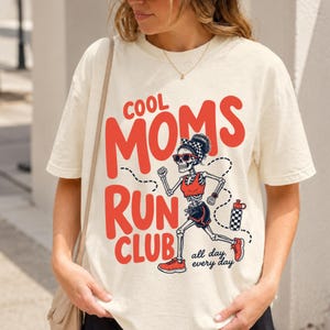 May include: A cream-colored short-sleeve t-shirt featuring a graphic of a skeleton running. The text reads COOL MOMS RUN CLUB and all day, every day. The skeleton wears a red top, dark shorts, red sneakers, sunglasses, and a checkered headband.