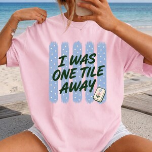May include: A light pink t-shirt featuring a graphic design of five vertical light blue stripes with white polka dots. The text "I WAS ONE TILE AWAY" is printed in dark green letters, accompanied by a small illustration of a mahjong tile.