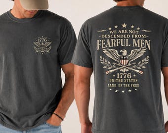 Peut inclure: T-shirts gris foncé avec des motifs patriotiques. L'un affiche "1776" et un emblème d'aigle. L'autre affiche "We Are Not Descended From Fearful Men" avec un aigle, un drapeau américain et le texte "1776". Les deux sont à manches courtes.