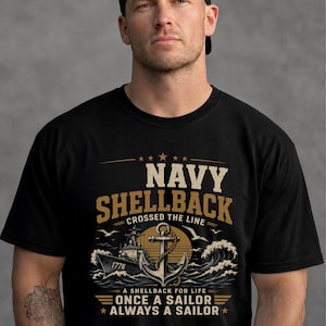 Può includere: Una t-shirt nera con una stampa grafica che riporta il testo NAVY SHELLBACK CROSSED THE LINE e A SHELLBACK FOR LIFE ONCE A SAILOR ALWAYS A SAILOR. Il design include una nave militare con la scritta 1776, un'ancora, onde e uccelli.