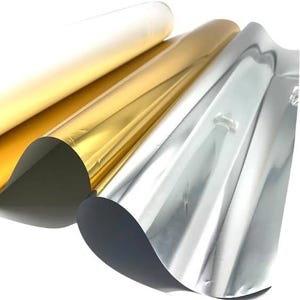 May include: Three sheets of metallic foil paper arranged in a fan-like display. The sheets feature a smooth, reflective surface in distinct colors: a matte white, a vibrant gold, and a bright silver, each showing gentle curves and folds.