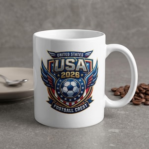 Pode incluir: Uma caneca de cerâmica branca com um gráfico colorido de uma bola de futebol centralizada num design de escudo. O texto diz UNITED STATES USA 2026 FOOTBALL CREST. O emblema inclui asas azuis, riscas vermelhas e brancas e estrelas douradas num fundo azul escuro.