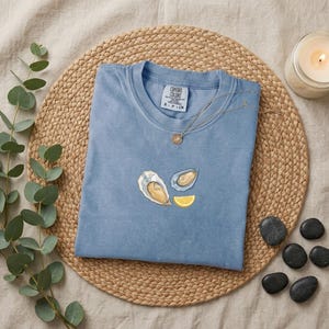 May include: A dusty blue t-shirt featuring an embroidered graphic of two oysters and a yellow lemon slice. The inner neck label displays the text Comfort Colors. A delicate gold-toned chain necklace rests on the collar of the shirt.