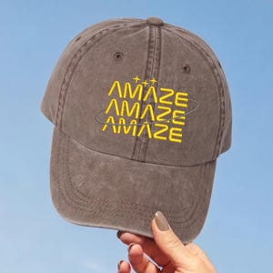 May include: A brown baseball cap with a distressed finish featuring the word AMAZE embroidered three times in yellow, stacked vertically. A thin yellow circular line encircles the text, and three small yellow four-pointed stars sit above.