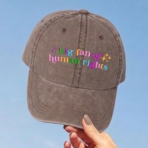 Könnte beinhalten: Eine braune Baseballkappe mit verwaschenem Finish und dem gestickten Text "big fan of human rights" in einer mehrfarbigen Schriftart. Das Design enthält kleine Sternsymbole und ist auf dem Vorderteil der sechsteiligen Kappe zentriert.