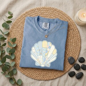 May include: A blue t-shirt featuring a large, central graphic of a seashell composed of various patchwork patterns in shades of light blue, yellow, and white. The shirt has a crew neckline and a Comfort Colors brand label at the inner collar.