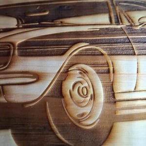 May include: A detailed wood carving of a classic car, showcasing the side profile. The artwork highlights the car's wheel, body, and windows, with intricate details. The wood has a natural, light brown color, and the carving is done with precision.