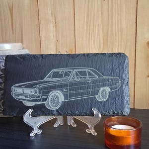 May include: A rectangular black slate sign featuring a detailed etched outline of a vintage Dodge Dart car. The car is depicted in profile, showcasing its classic design. The sign is supported by a clear stand.