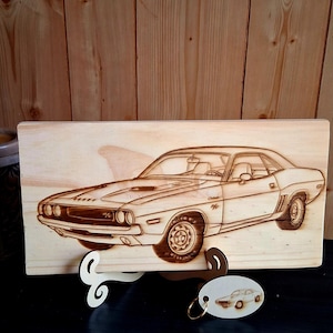 May include: A wooden plaque featuring a detailed pyrography image of a classic Dodge Challenger. The car is depicted in profile, with intricate details of the body, wheels, and interior. A small keychain with a car image hangs below.