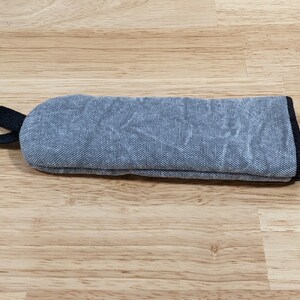 May include: A rectangular, padded fabric sleeve in a textured, mottled blue-grey color. The sleeve features a black fabric loop attached to one end and is finished with a black fabric trim along the edges, providing a structured, protective case.