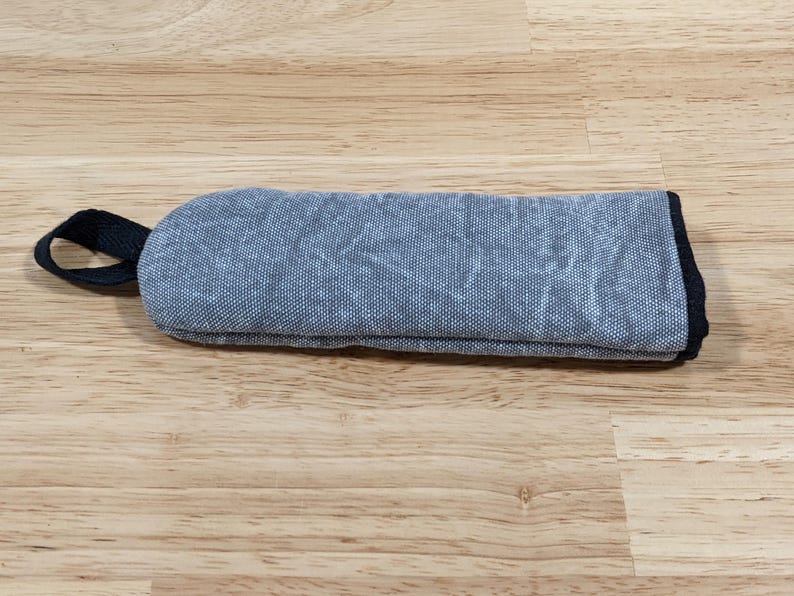 May include: A rectangular, padded fabric sleeve in a textured, mottled blue-grey color. The sleeve features a black fabric loop attached to one end and is finished with a black fabric trim along the edges, providing a structured, protective case.
