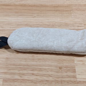 May include: A rectangular oven mitt featuring a light gray, textured fabric exterior. The mitt includes a dark navy blue fabric loop attached to one side for hanging and a matching dark navy blue trim along the open end of the mitt.