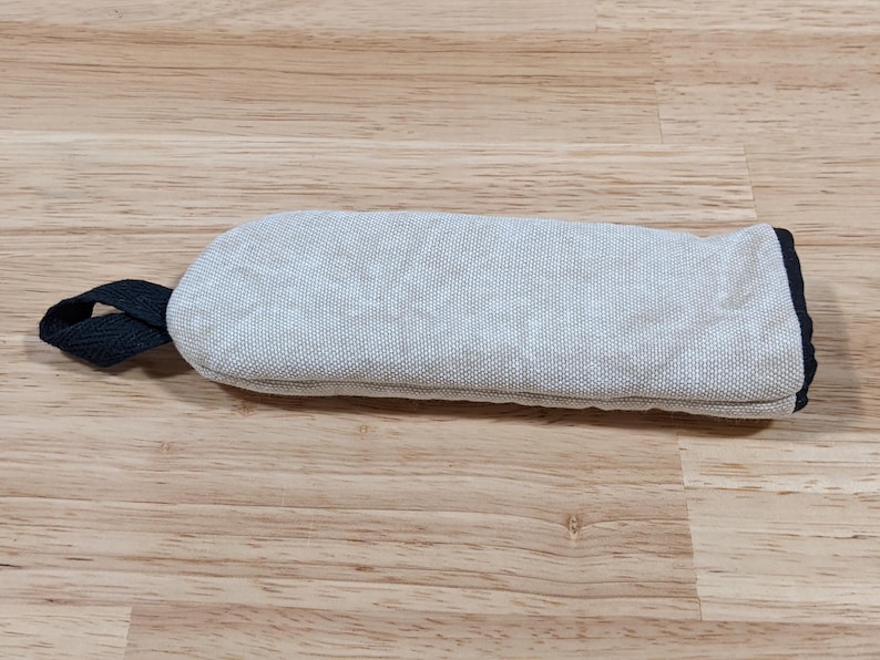 May include: A rectangular oven mitt featuring a light gray, textured fabric exterior. The mitt includes a dark navy blue fabric loop attached to one side for hanging and a matching dark navy blue trim along the open end of the mitt.