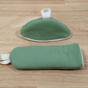 May include: A sage green fabric oven mitt and a matching sage green fabric pot holder. Both items feature a white fabric trim along the edges and a white fabric loop for hanging. The pot holder is semi-circular and the oven mitt is rectangular.