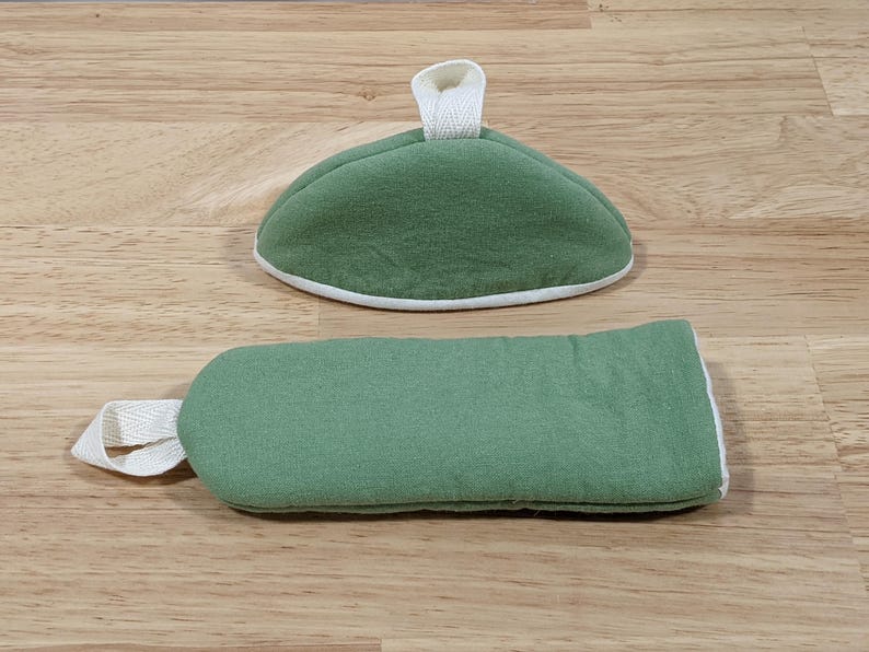 May include: A sage green fabric oven mitt and a matching sage green fabric pot holder. Both items feature a white fabric trim along the edges and a white fabric loop for hanging. The pot holder is semi-circular and the oven mitt is rectangular.