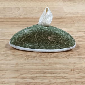 May include: A dome-shaped fabric tea cozy featuring an olive green background with a white botanical print of leaves and flowers. The item has a white fabric trim along the bottom edge and a white fabric loop handle attached to the top center.