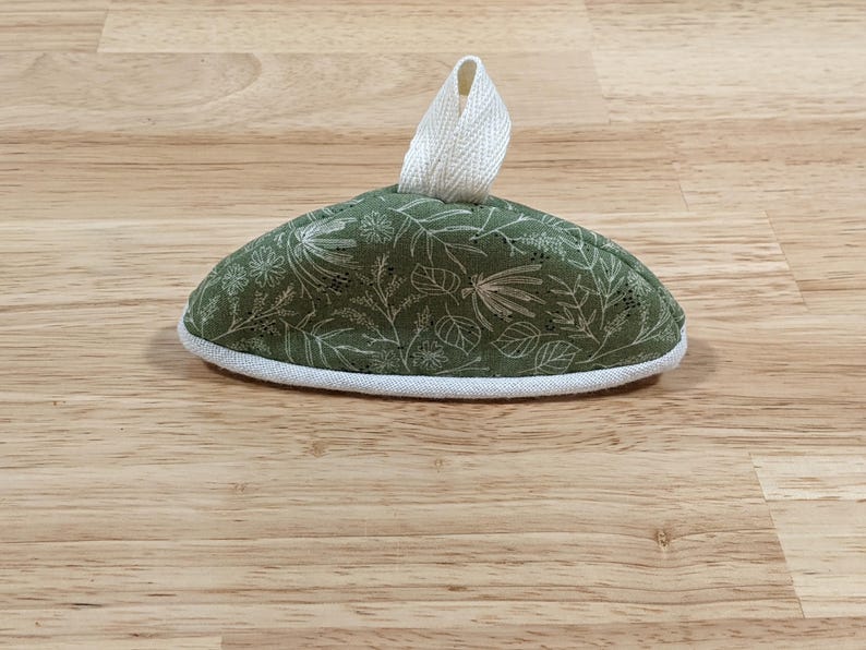 May include: A dome-shaped fabric tea cozy featuring an olive green background with a white botanical print of leaves and flowers. The item has a white fabric trim along the bottom edge and a white fabric loop handle attached to the top center.