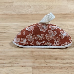 May include: A dome-shaped fabric pot handle cover featuring a rust-colored background with a white floral pattern. The item includes a white herringbone-patterned fabric loop at the top and is finished with a white fabric trim along the bottom edge.